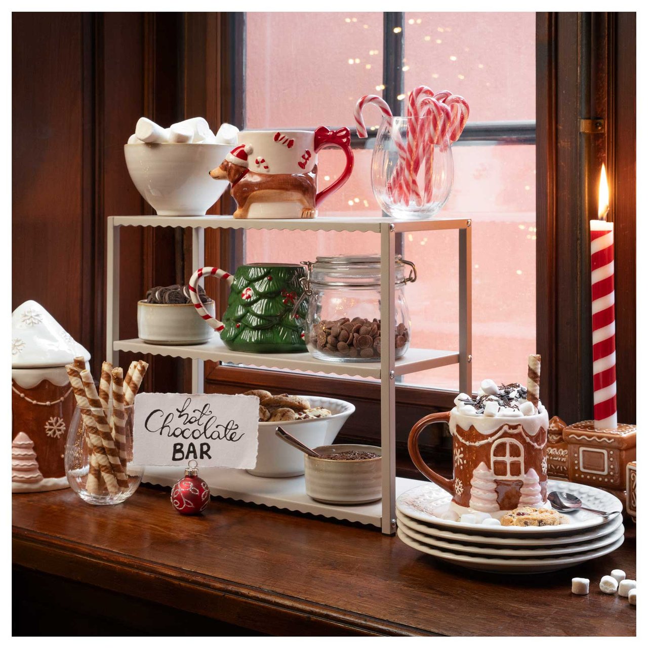 En hot chocolate bar uppdukad i ett fönster med julmuggar, marshmallows, polkagrisar och tända ljus.