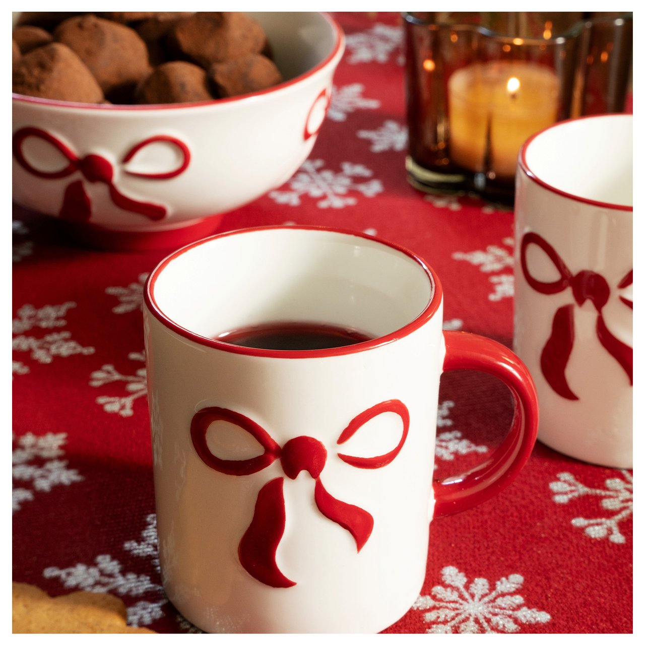 Julmugg med röd rosett på röd duk med snöflingor, skål med chokladtryfflar och tänd ljuslykta.