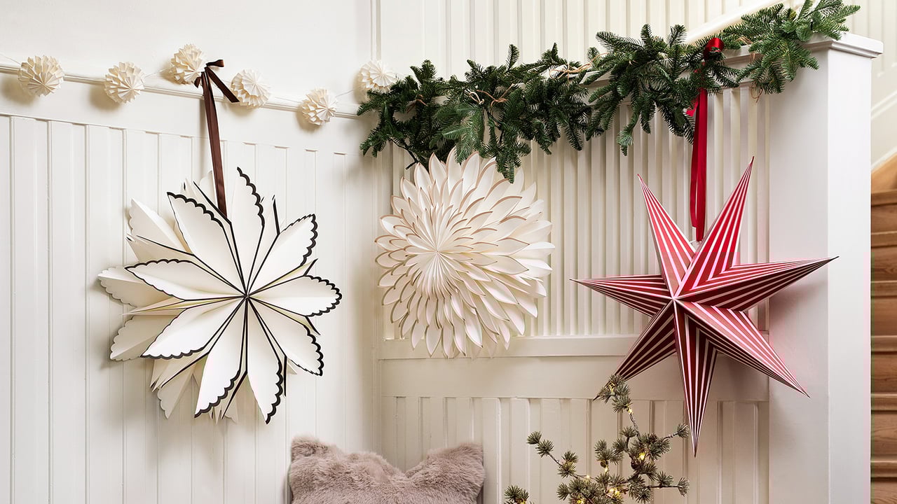 Drei Papiersterne in Weiß und Rot hängen an einer mit Tannengirlande und Lichterkette geschmückten Wand.