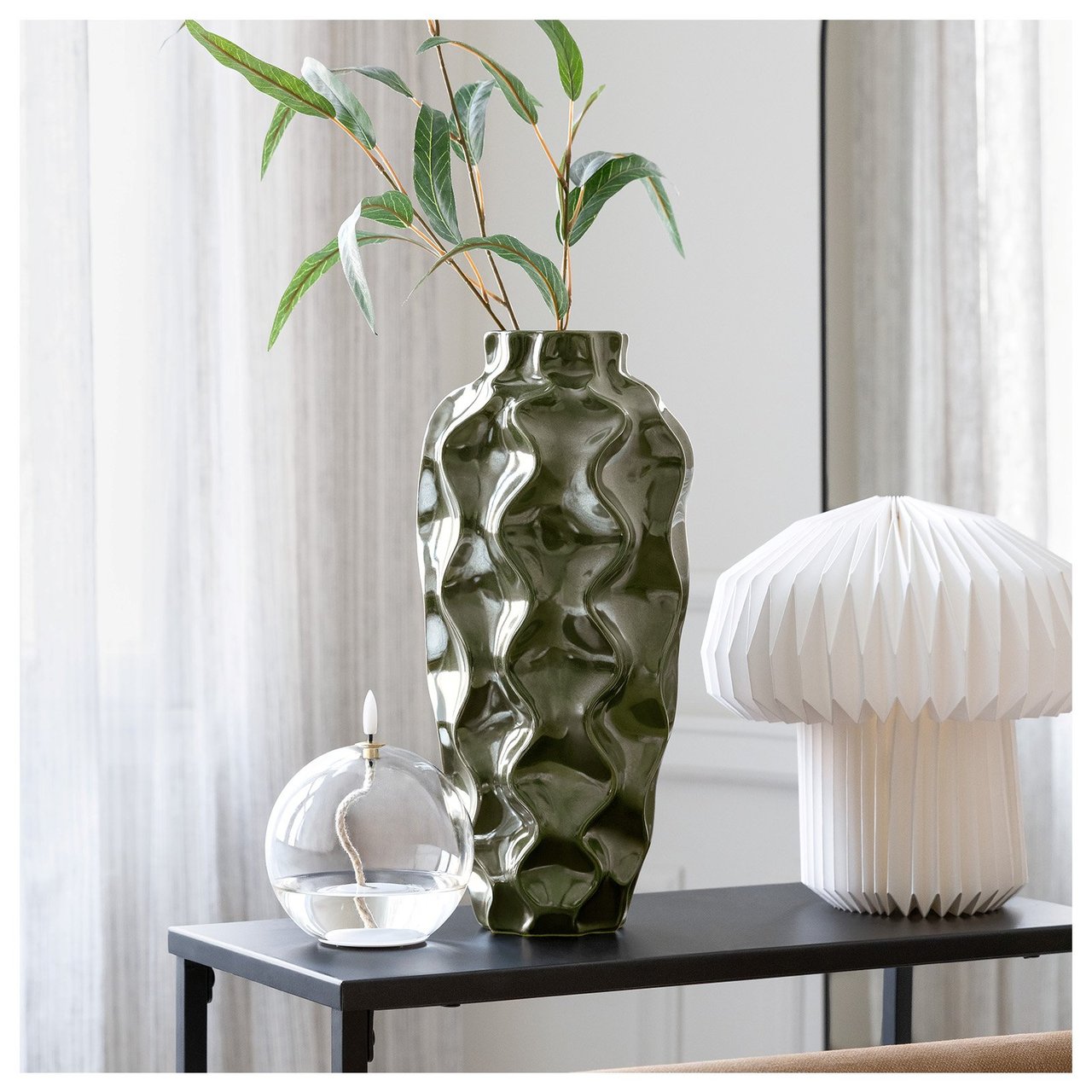 En høy, grønn vase står sammen med en hvit lampe og en lyslykt på et sidebord.