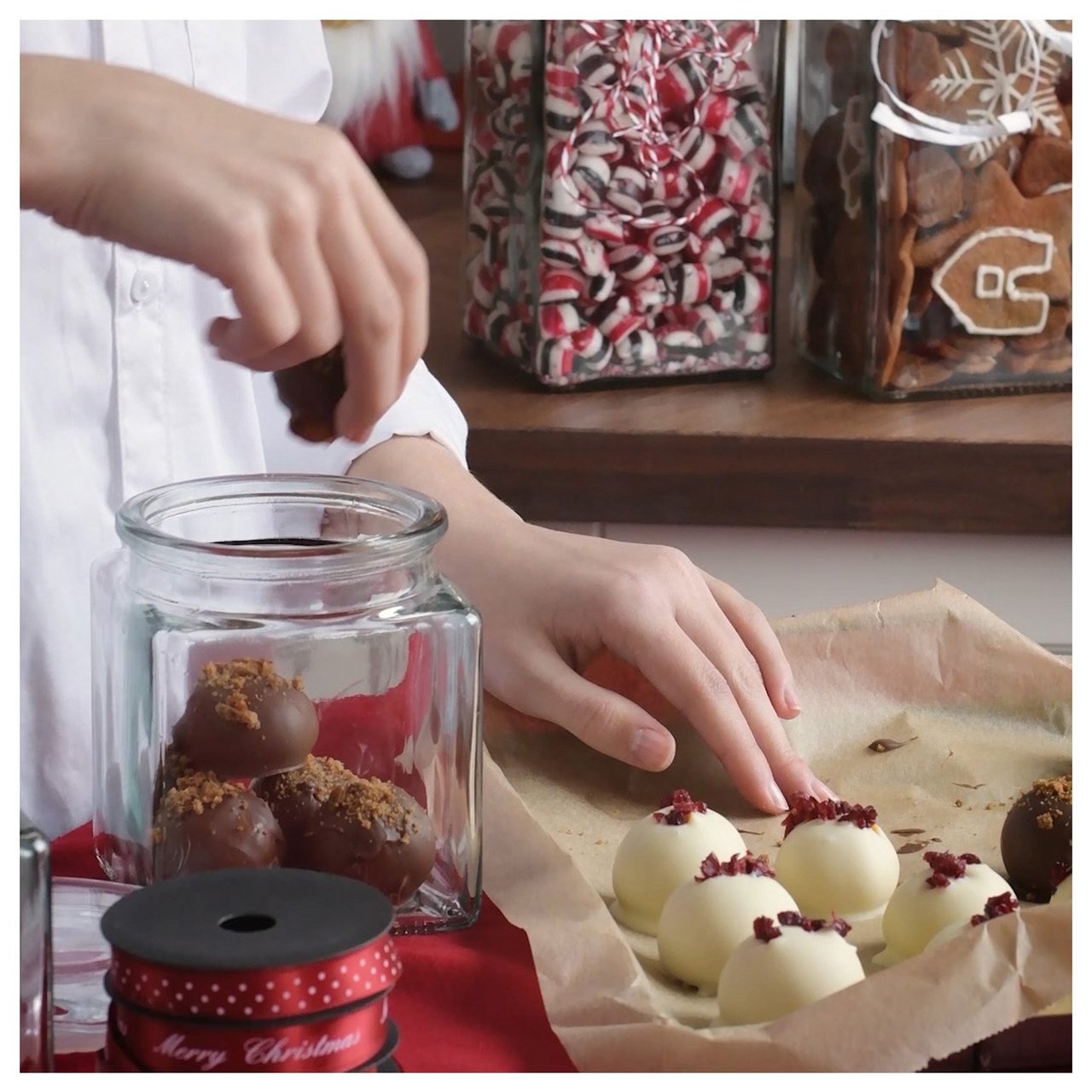 Käsi laittaa suklaapaloja lasipurkkiin ja vieressä on valkoisia konvehteja leivinpaperilla.