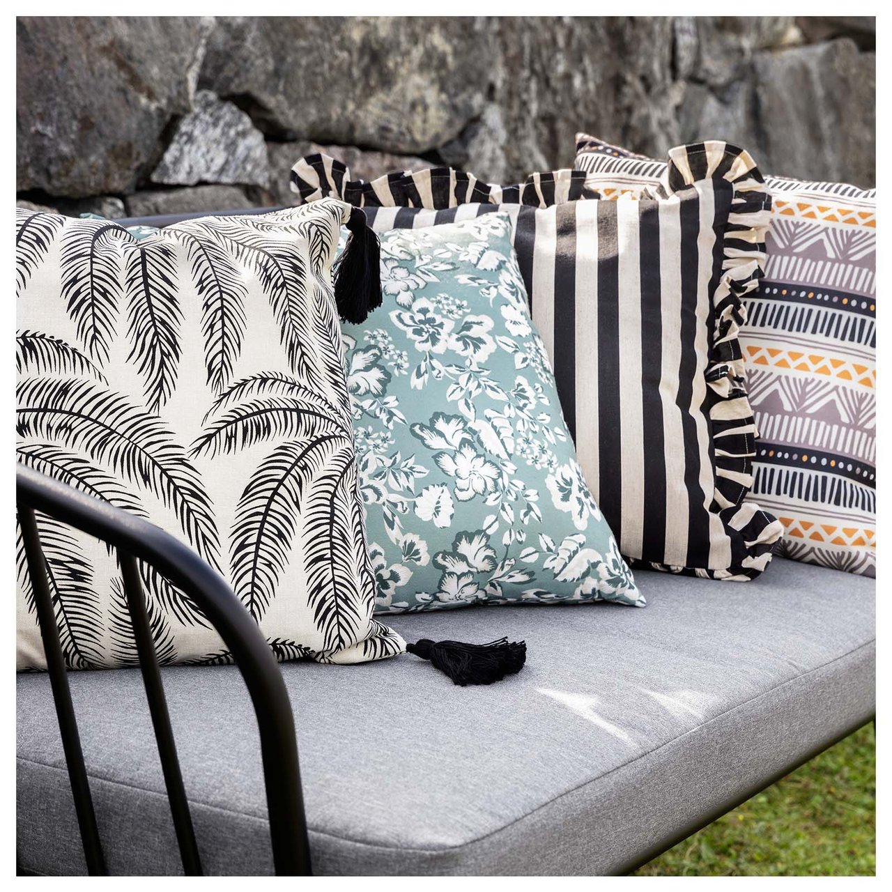 Lounge-Sofa in Grau mit vier bunten Kissen in verschiedenen Mustern, das vor einer Steinmauer steht.