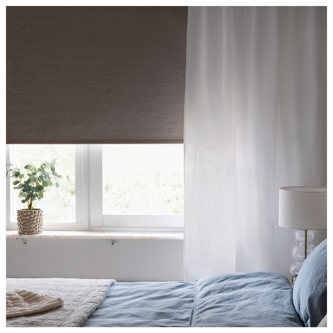 Makuuhuone, jossa on sänky, verhot ja rullaverho ikkunan edessä sekä pieni kasvi ikkunalaudalla.