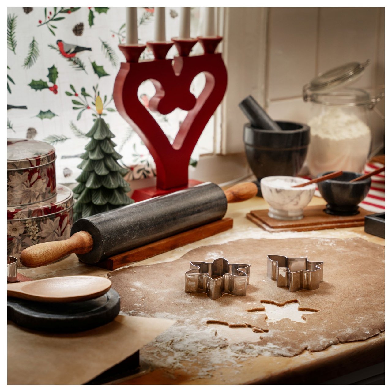 Keittiössä leivotaan piparkakkuja, ja siellä on joulukoristeita, kaulin ja muotteja jauhotetulla pöydällä.