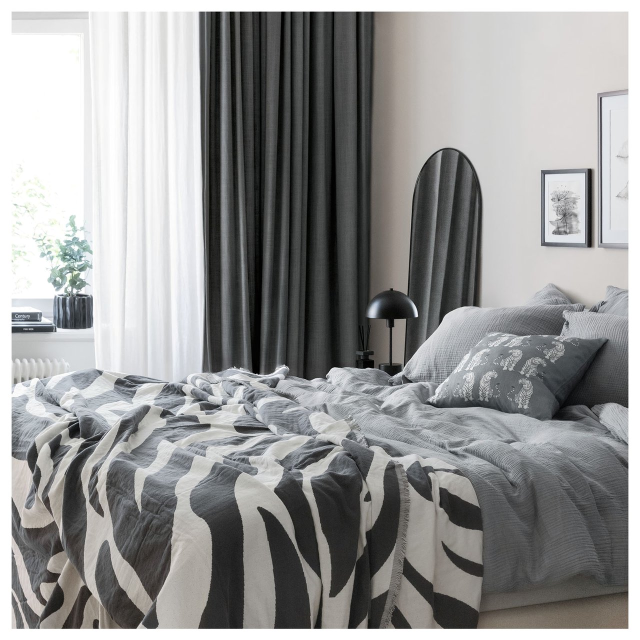 Schlafzimmer mit grauer Bettwäsche und einem dünnen weißen sowie einem dicken dunkelgrauen Vorhang zusammen.