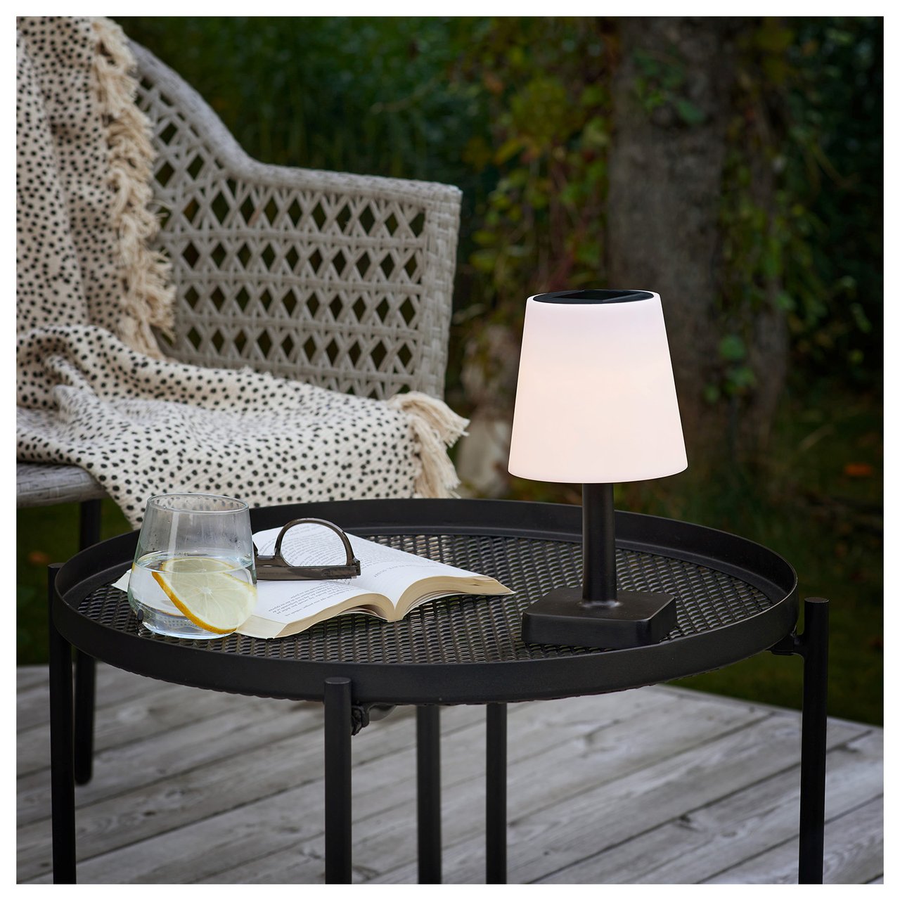 Kleiner Tisch auf der Terrasse bei Abenddämmerung mit Solarlampe, neben einem Rattanstuhl mit Decke.