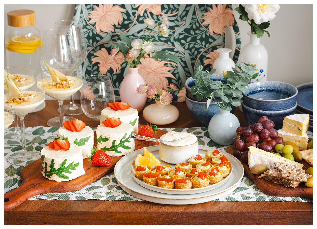 Festlich gedeckter Tisch mit Desserts, Schnittchen, Käseplatte und Getränken in dekorierter Umgebung.