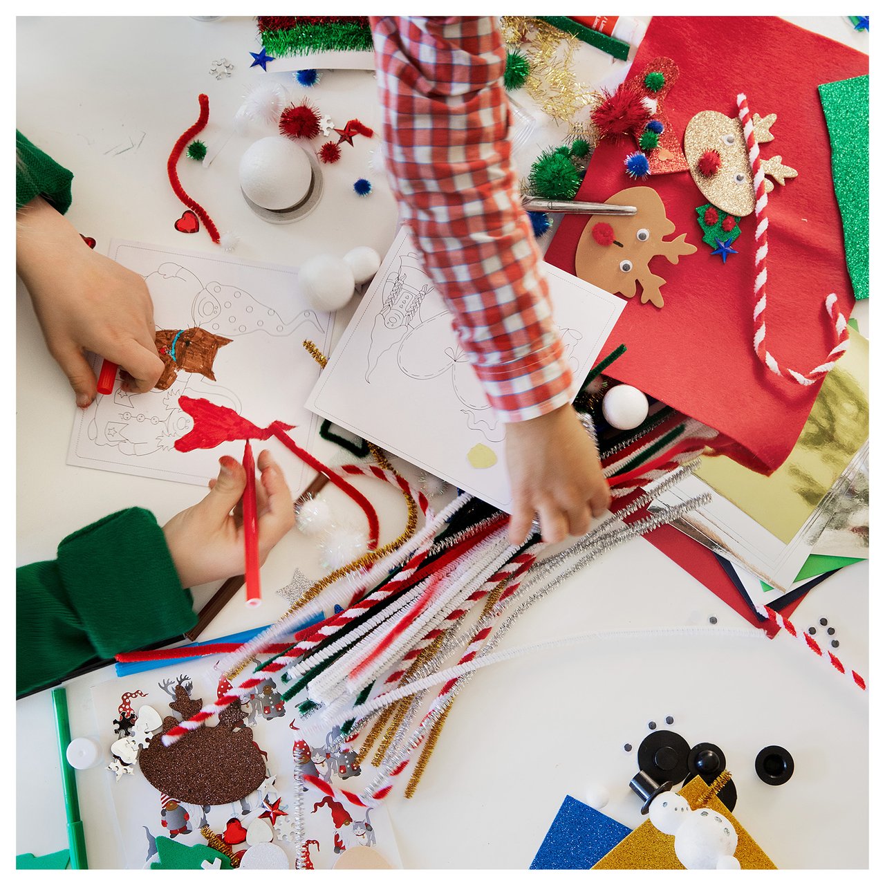 Lasten kädet, jotka askartelevat jouluaiheisia asioita pöydällä, joka on täynnä värikkäitä tarvikkeita.