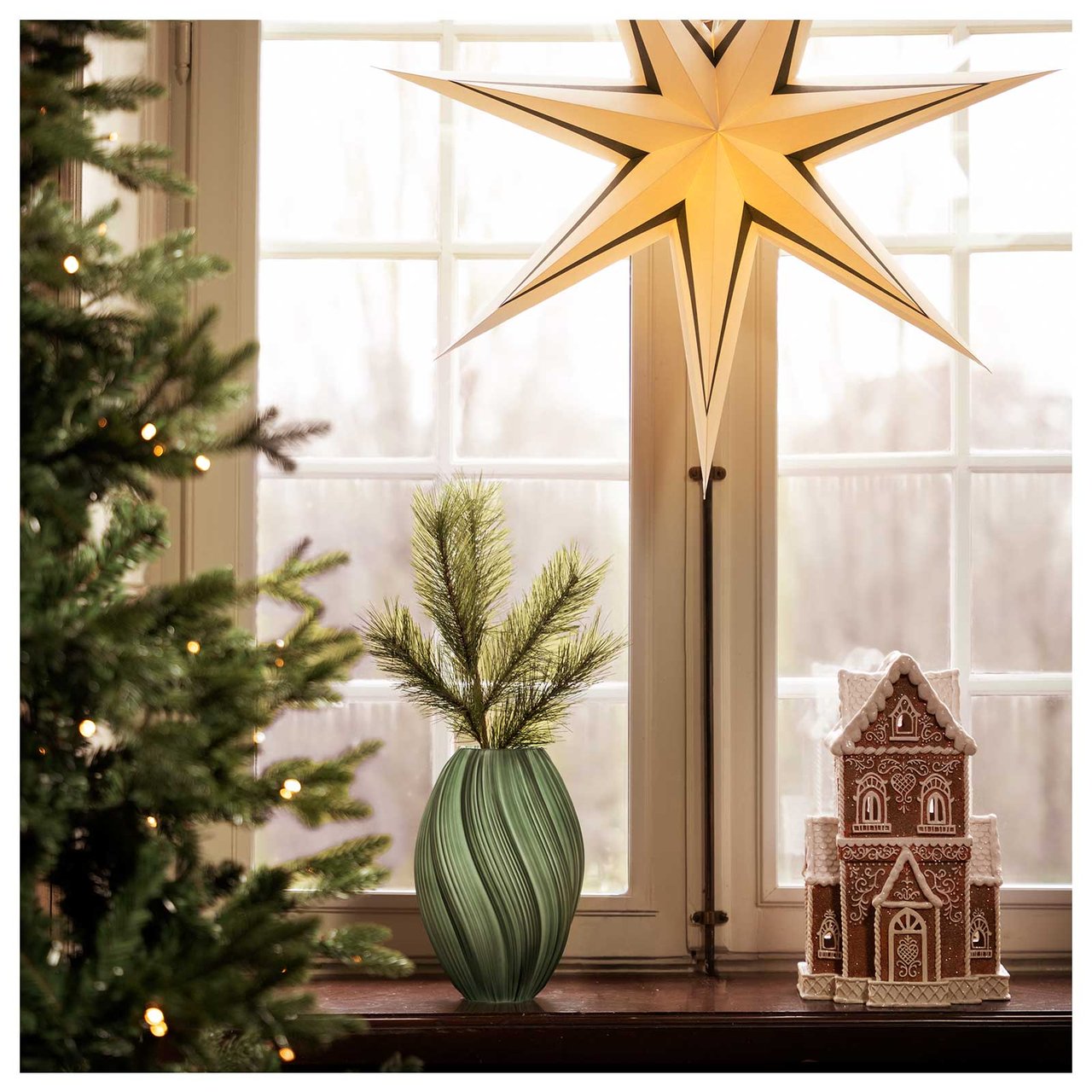 En vit adventsstjärna med grön rand hänger i ett fönster. En vas med grankvist och ett pepparkakshus står på fönsterbrädet.