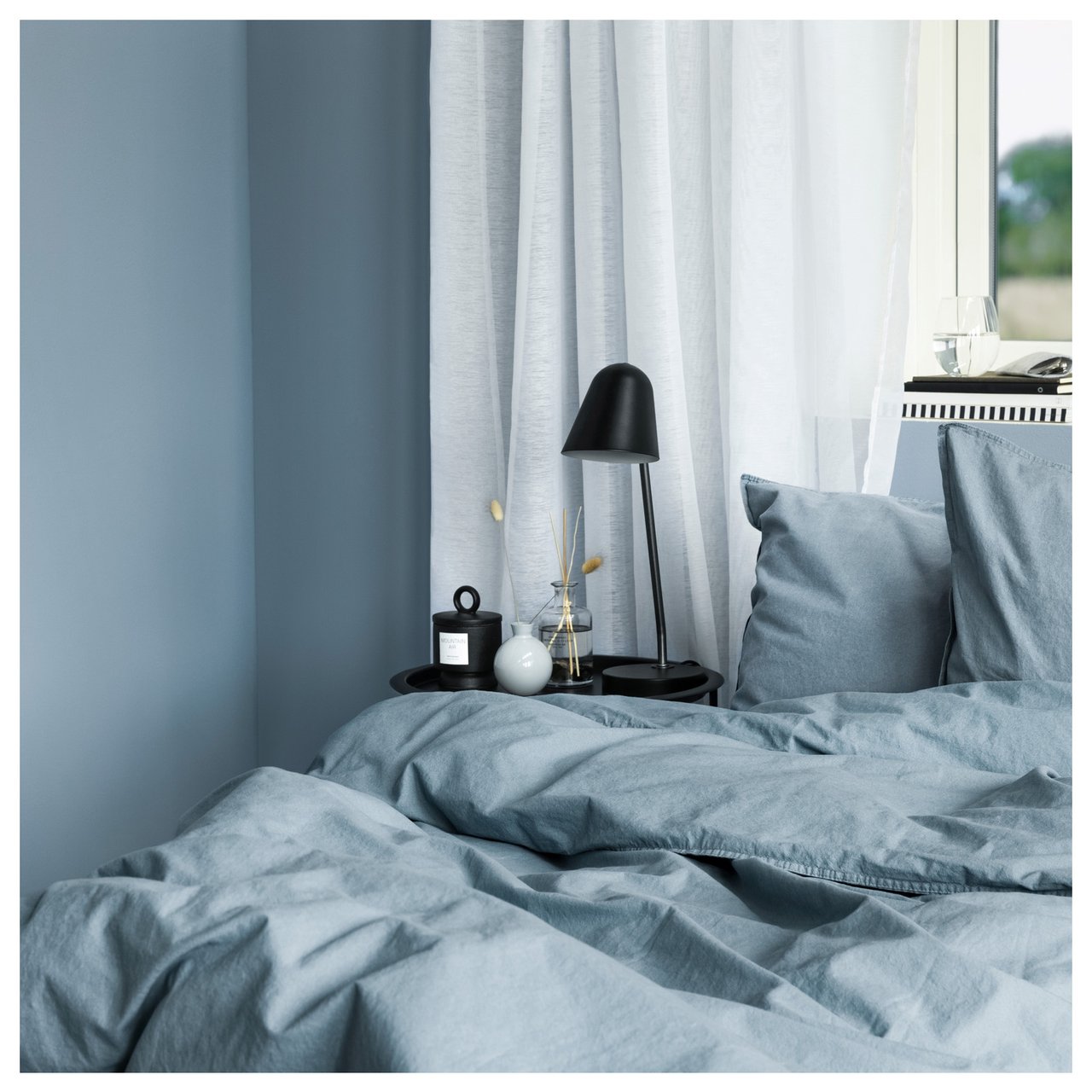 Schlafzimmer mit Betttextilien und Wandfarbe in Blautönen vor einem Fenster mit durchsichtigen Vorhängen.