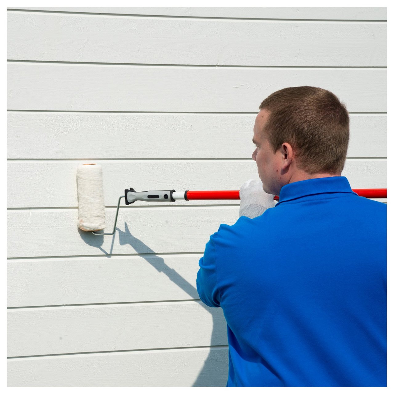 Man med blå t-shirt målar en vit husfasad med roller som har ett förlängningsskaft.