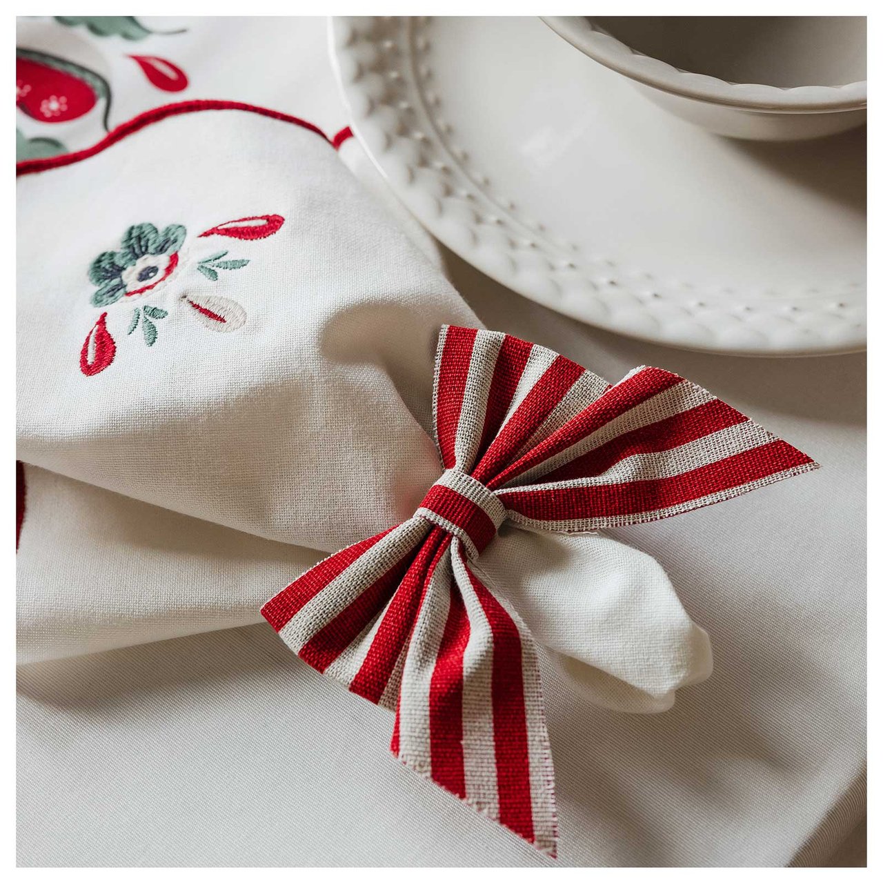 En broderad tygservett med en rödvitrandig rosett ligger intill en vit tallrik på ett bord med julduk.