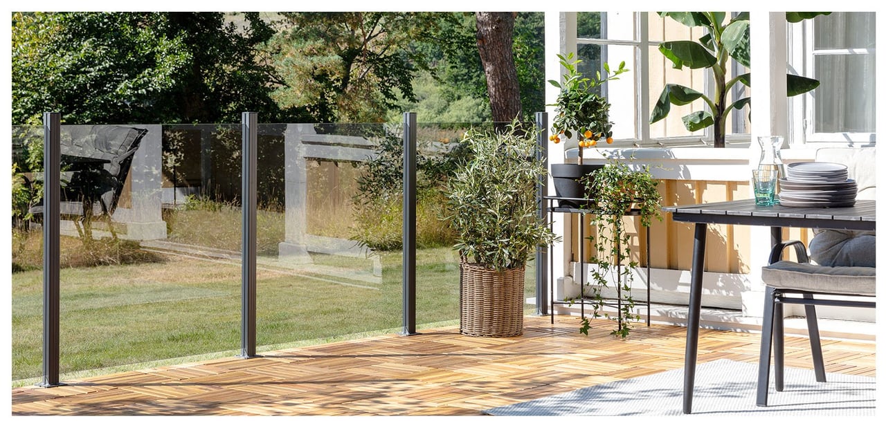 Solig altan med trätrall som ramas in av glasräcken mot trädgård.