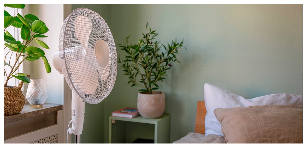 Golvfläkt placerad i ett sovrum med växter, säng och ett nattduksbord.