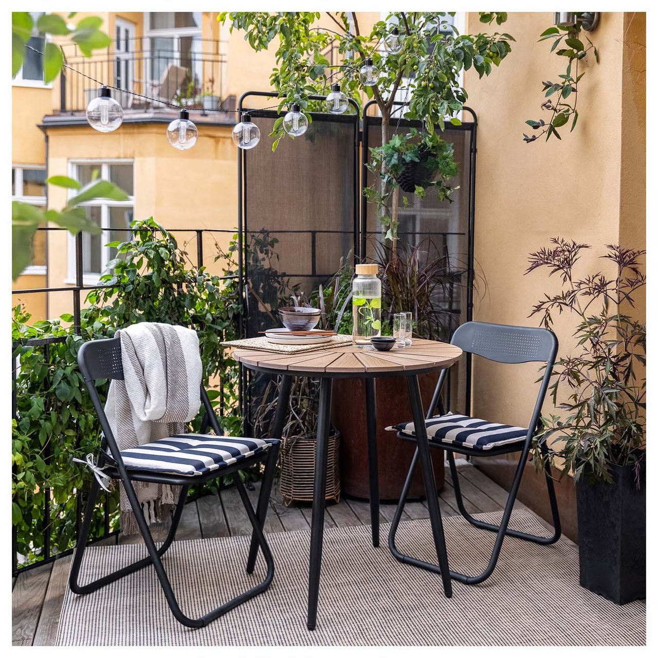 Kompakte Balkonmöbel mit zwei Klappstühlen und einem Tisch aus schwarzem Stahl stehen auf einem Balkon mit Sichtschutz.