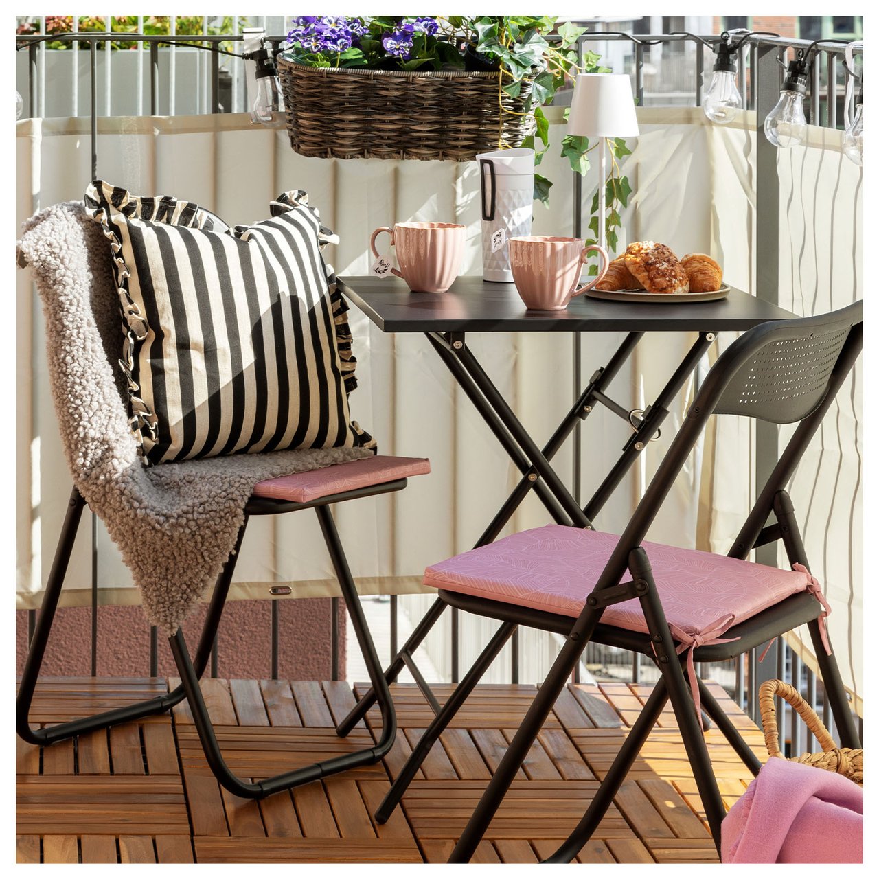 Kompakte Balkonmöbel mit zwei Klappstühlen und einem Tisch aus schwarzem Stahl stehen auf einem Balkon mit Sichtschutz.