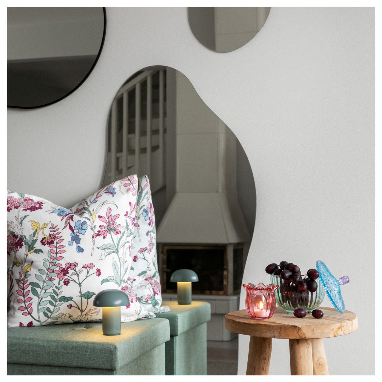 Erilaisia peilejä seinällä, rahi, pienet pöytävalaisimet ja kukkakuvioinen tyyny olohuoneessa.