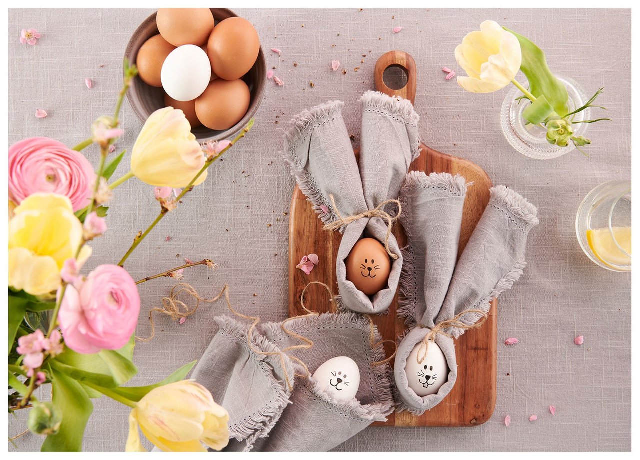 Ostereier mit Hasengesichtern, eingeschlagen in graue Servietten, umgeben von Blumen und Dekorationen.