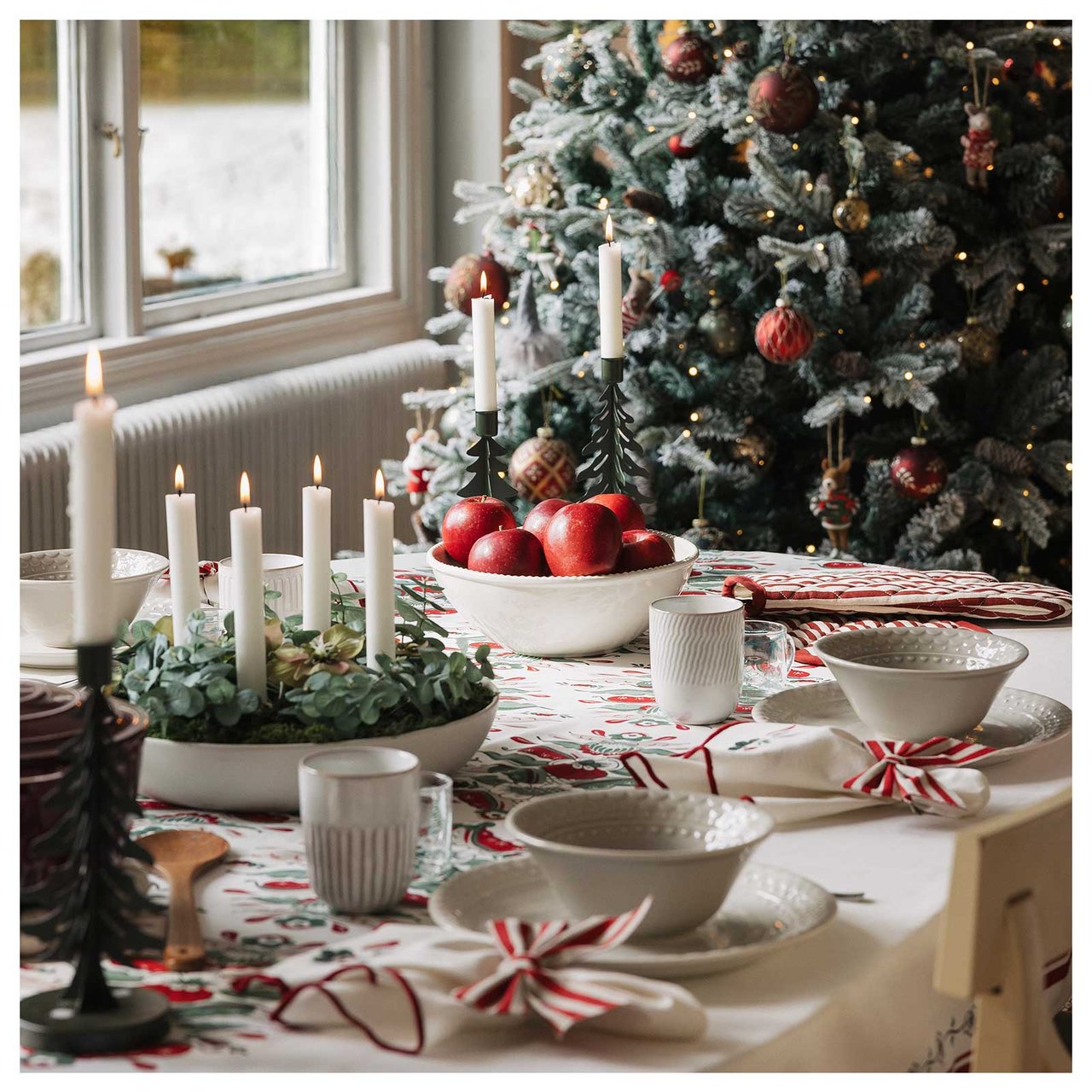 Ett juldukat bord i vitt och rött, med en julgran och en adventsstjärna i bakgrunden. 