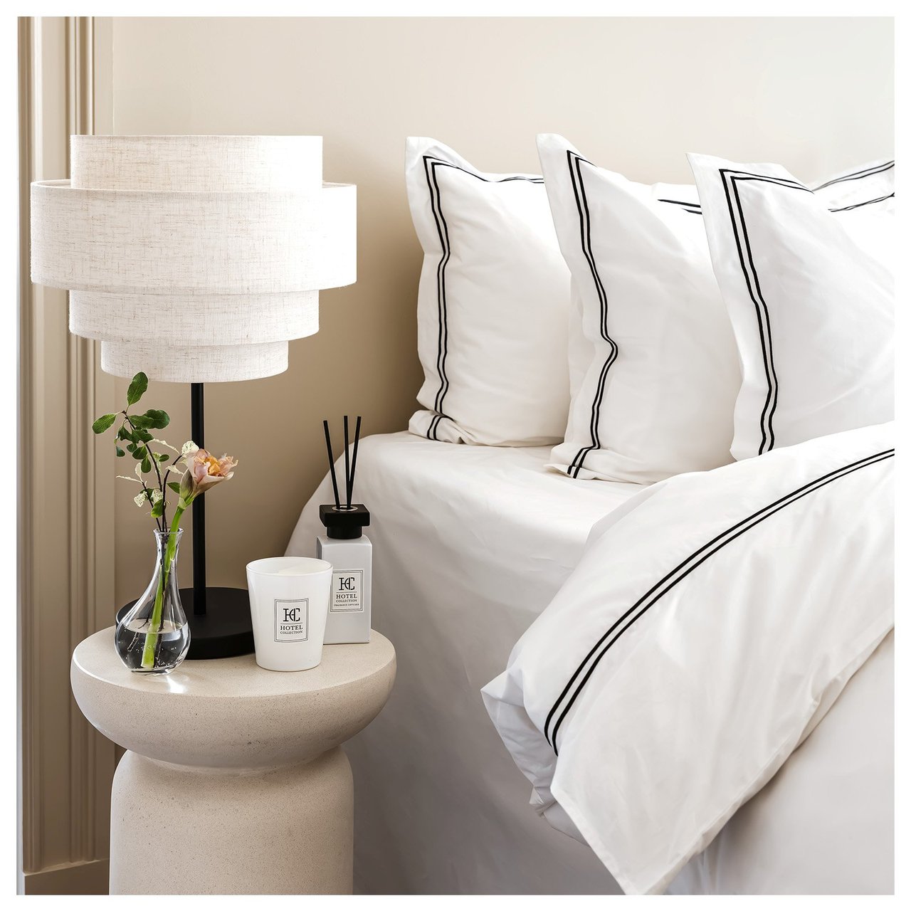 Nachttisch mit weißer Tischlampe, Duftkerze und Duftstäbchen. Bett mit weißer Bettwäsche mit schwarzen Details.
