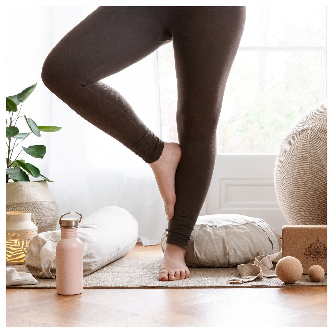 Person står i yogaposition i ett ljust rum med träningsutrustning, vattenflaska och kuddar.