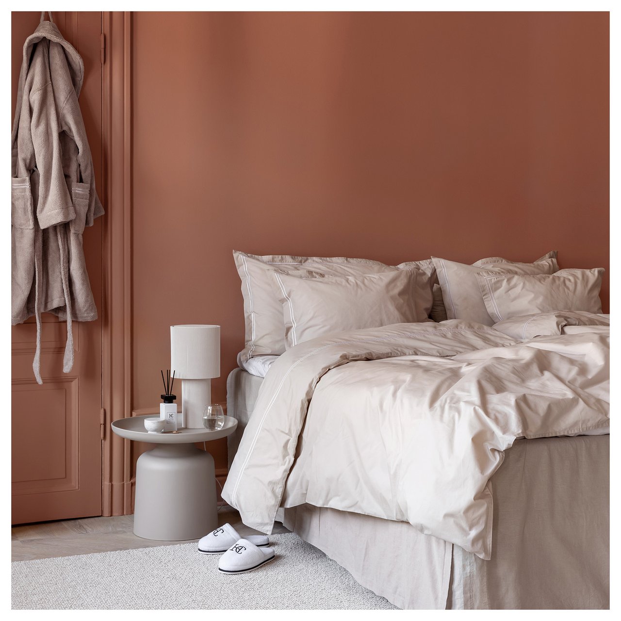 Soverom med dobbeltseng. Beige sengetøy med hvit, dekorativ rand. En beige morgenkåpe henger på døren.