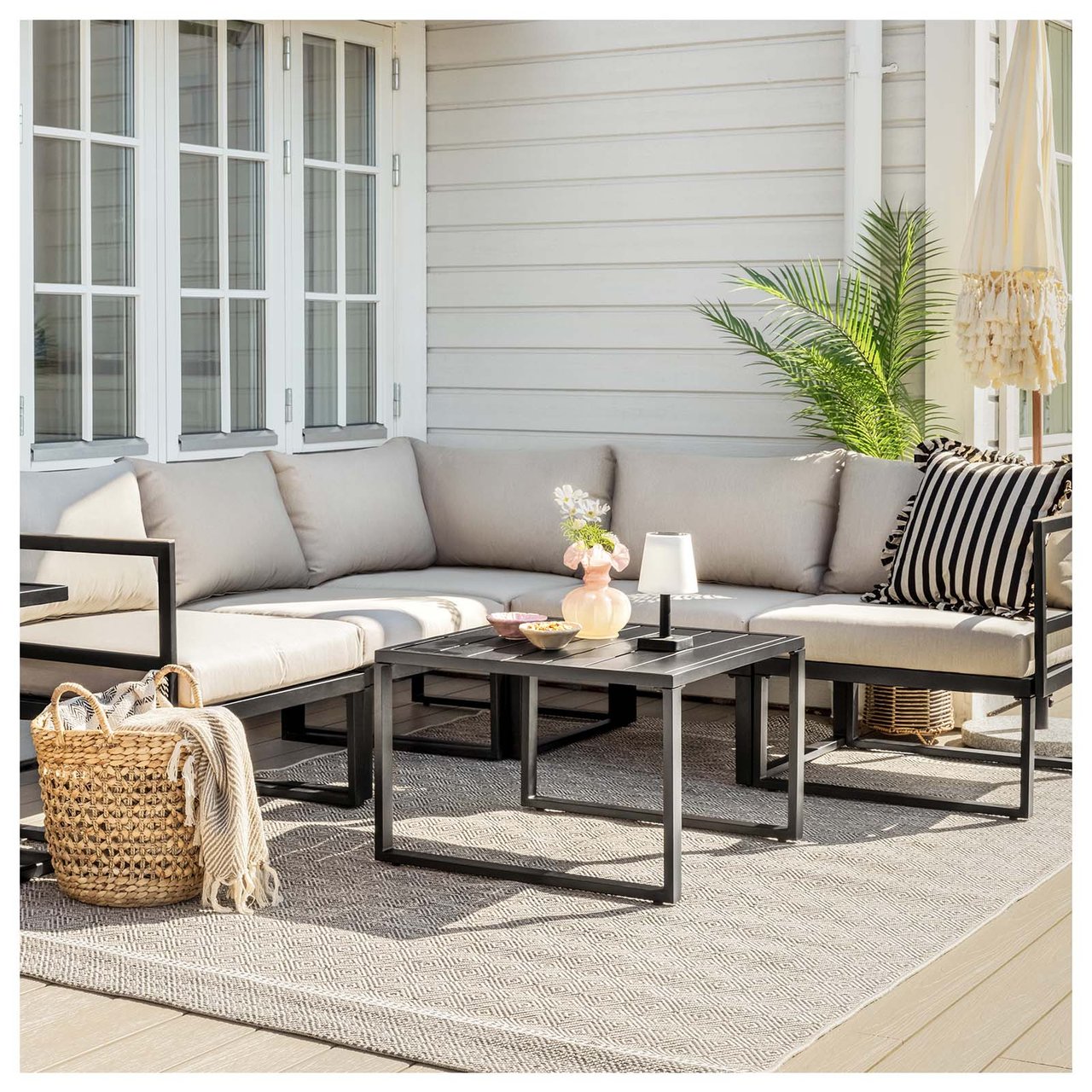 Großes Ecksofa mit beigen Sitzkissen auf einer Terrasse vor einer Hauswand. Das Sofa hat beige Sitzkissen, ein Couchtisch ist mit einer Solarlampe dekoriert.