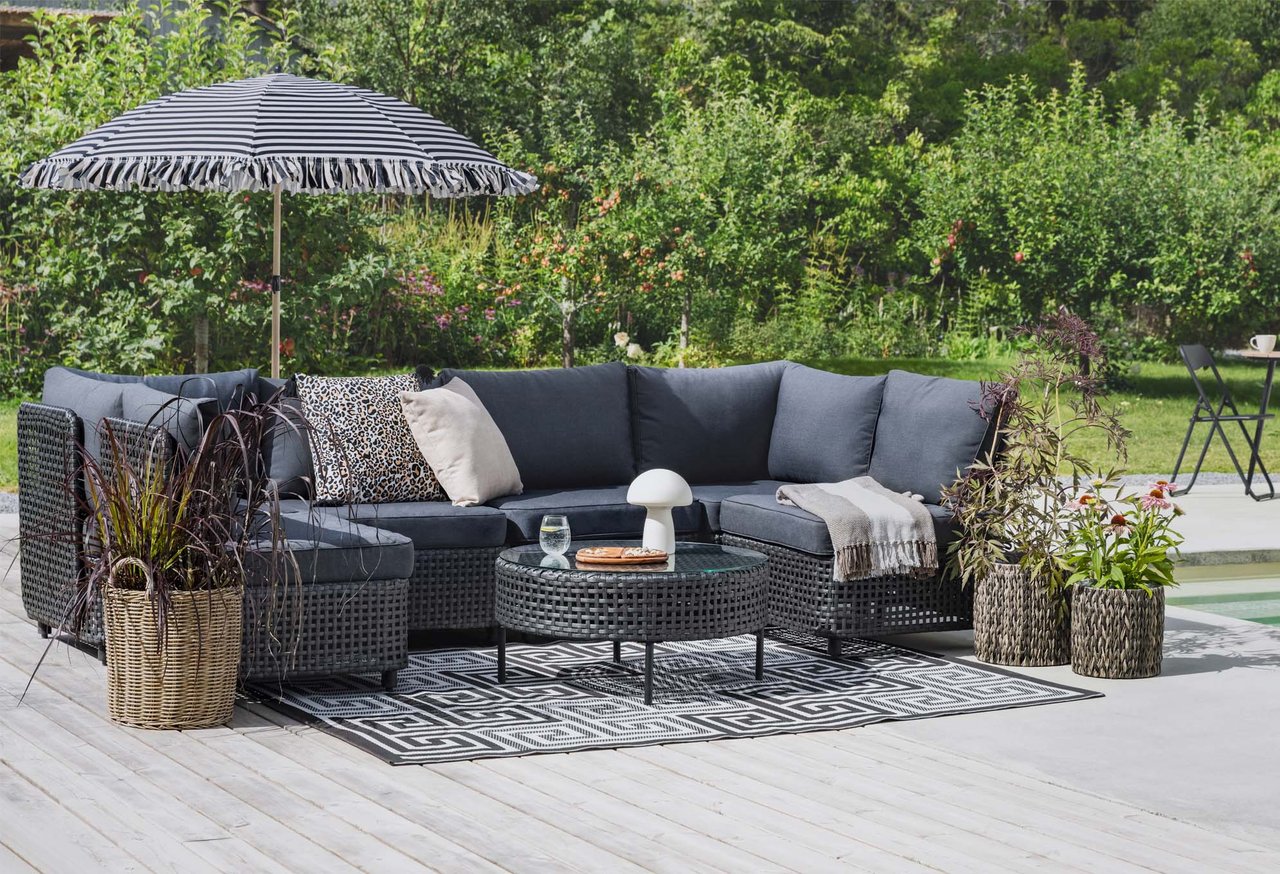 Gemütliche Terrasse mit grauem Loungesofa, Sonnenschirm, Tisch und Pflanzen im Garten.