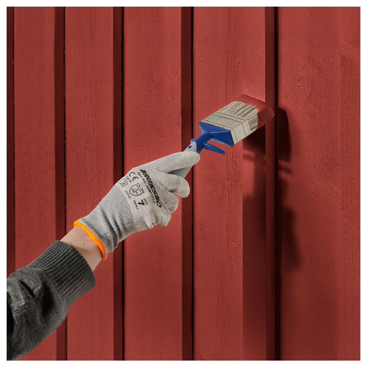 Hand med greppvänlig målarhandske målar en husfasad i röd färg med en bred pensel.