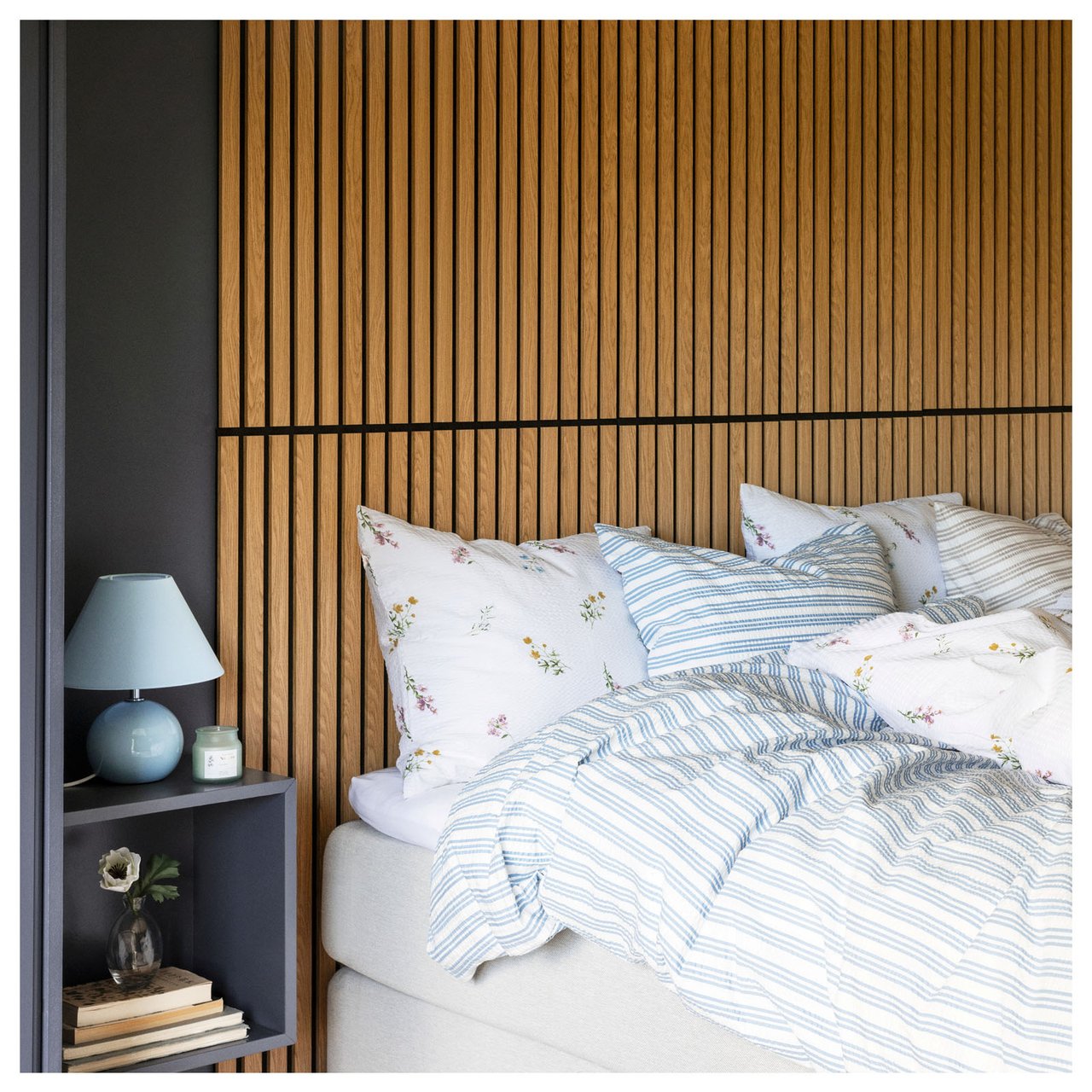 Das Bett ist mit heller Bettwäsche bezogen. Hinter dem Bett ist eine Akustikpaneele als Kopfteil montiert. Nachttisch mit Tischlampe auf der linken Seite.