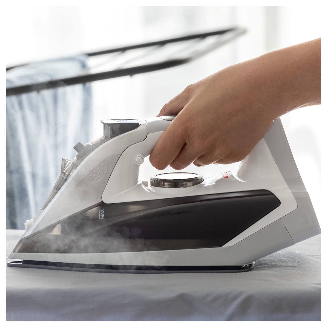 Hand stryker med ett strykjärn på en strykbräda framför ett torkställ.