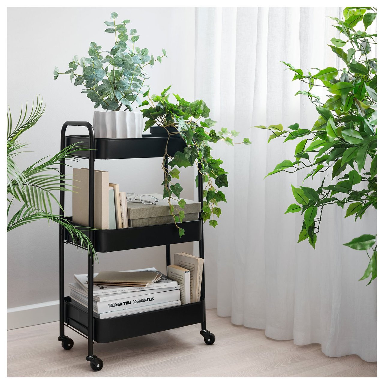 Ablagewagen mit Büchern und Pflanzen in einem hellen Raum mit Vorhängen und grünen Akzenten.