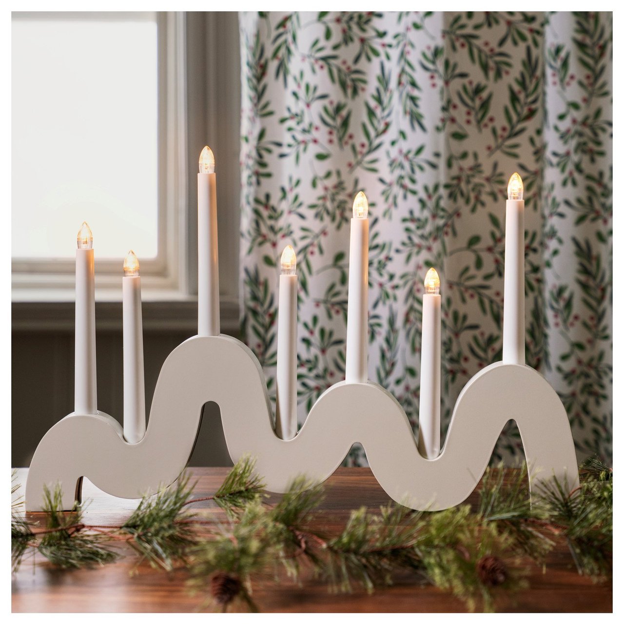 Moderni sähkökynttelikkö aaltoilevalla muodolla valaisee huonetta jouluisen verhon edessä.