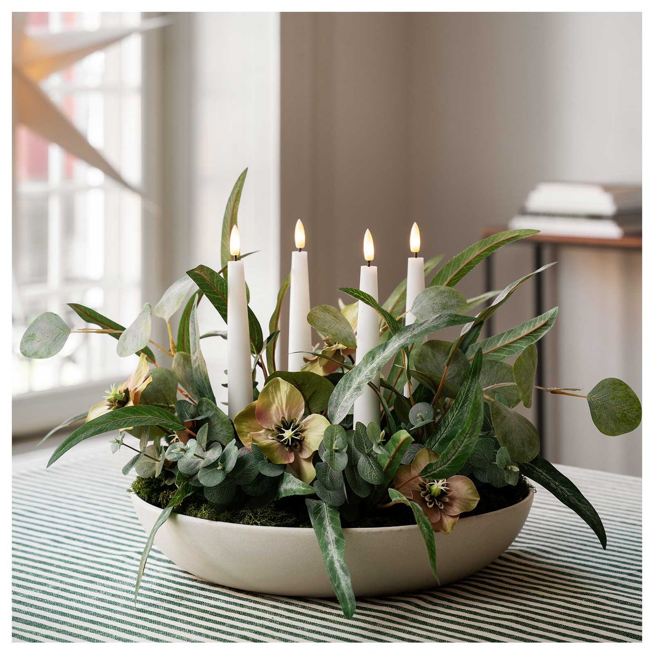 En vit adventsljusstake dekorerad med gröna kvistar och julrosor står på ett bord med grönvitrandig duk. 