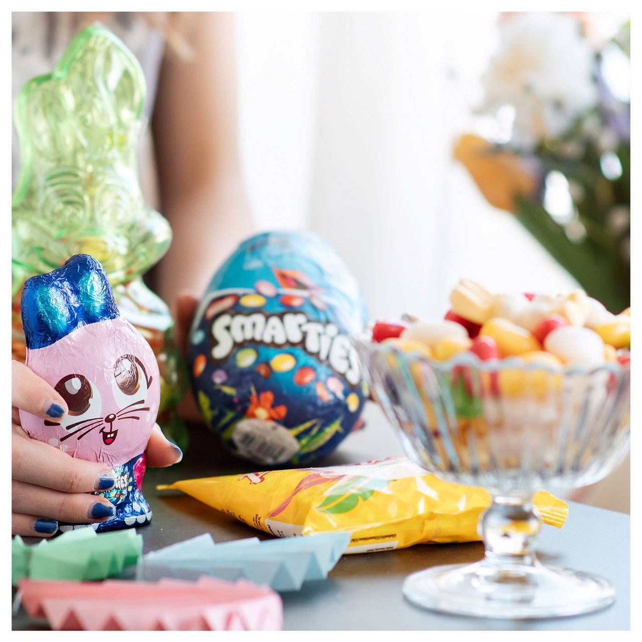 Bonbonschale aus Glas, Osterei und Schokohase auf einem Tisch in Nahaufnahme.