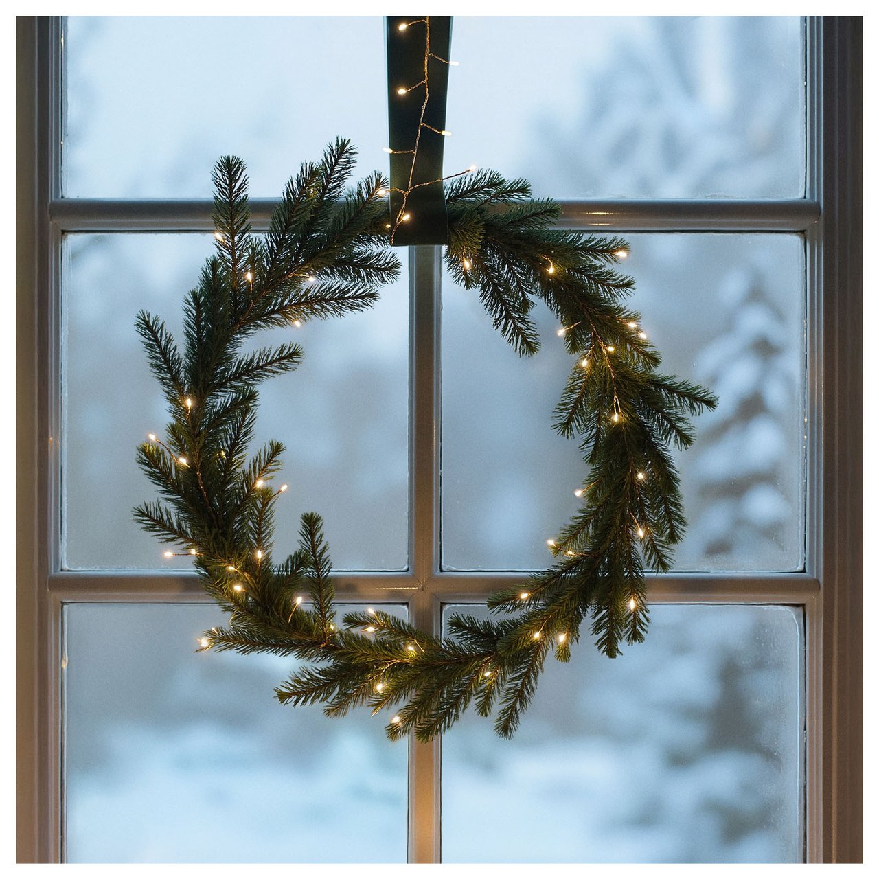 En krans av granris pyntad med en ljusslinga hänger i ett fönster.