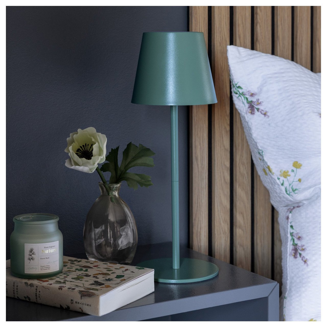 Yöpöytä, jolla on vihreä pöytävalaisin, kukkamaljakko, kirja ja tuoksukynttilä sijatun sängyn vieressä.