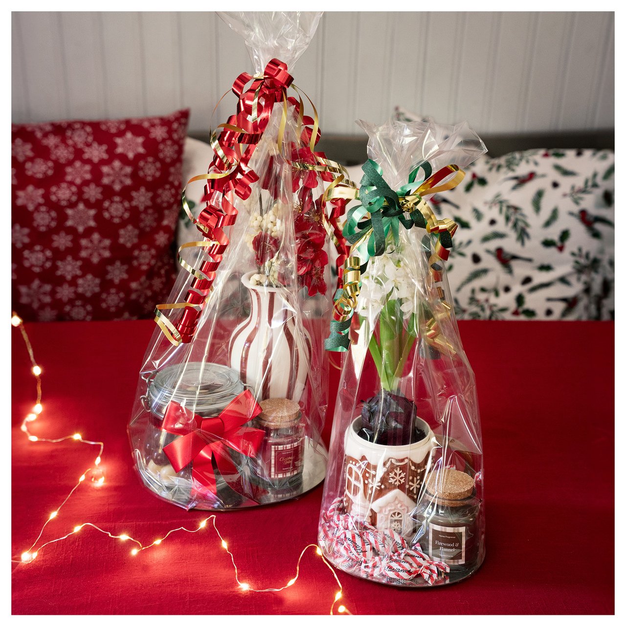 Kaksi sellofaaniin käärittyä joululahjakoria, joissa on koristeita, herkkuja ja lahjoja punaisella pöydällä.