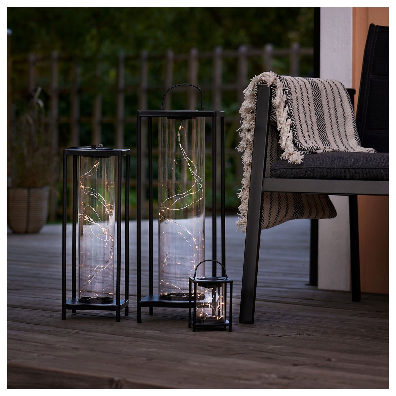 Del av altan där det står en stol med pläd i samt tre solcellslyktor i glas och metall med olika höjd.