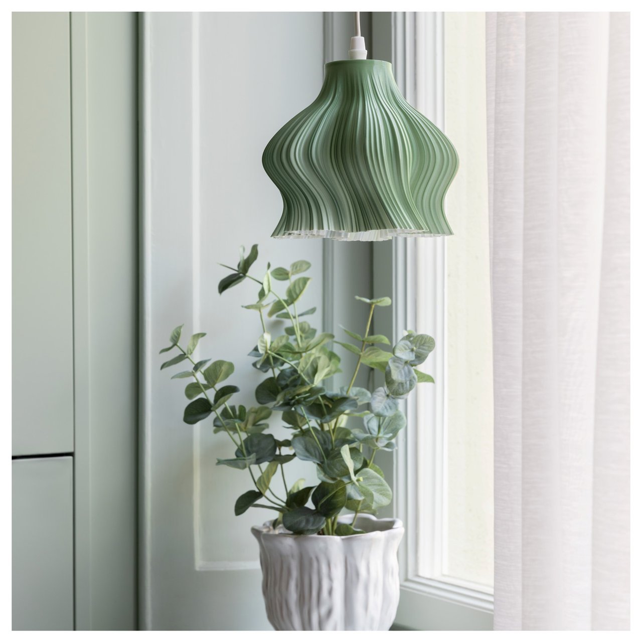 Taklampa med skulptural form hänger ovanför en krukväxt vid ett fönster.