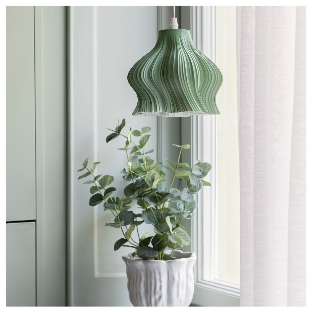Taklampa med skulptural form hänger ovanför en krukväxt vid ett fönster.