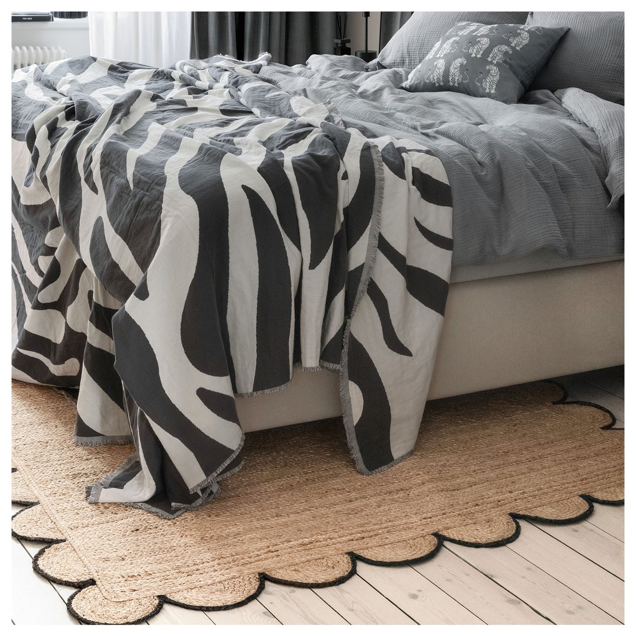 Soverom med grått sengetøy, sebramønstret sengeteppe og juteteppe med svart, bølgete kant.