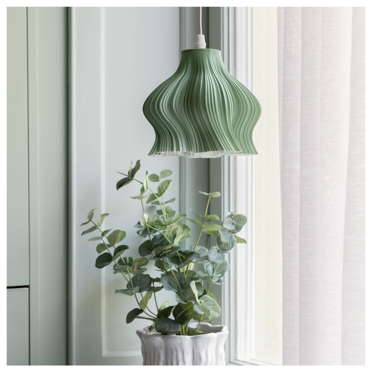 En liten grønn vinduslampe med bølgete former henger i et vindu. I vinduskarmen står det en grønn potteplante.