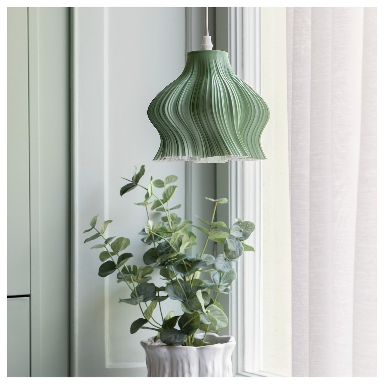 En liten grønn vinduslampe med bølgete former henger i et vindu. I vinduskarmen står det en grønn potteplante.