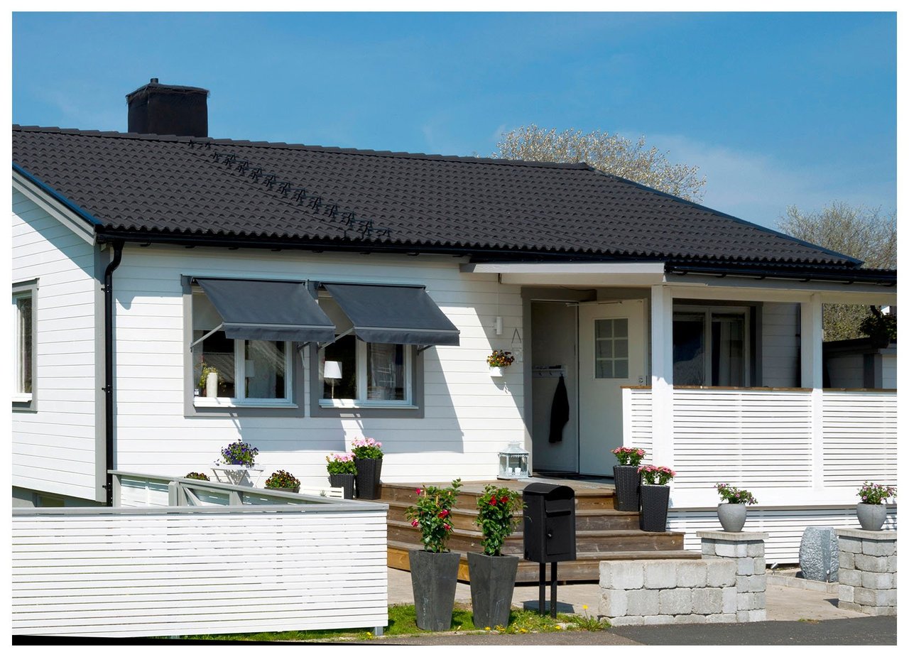 Vitmålad enplansvilla med gråa knutar, svart tak, mörkgrå fönstermarkiser och terrass.