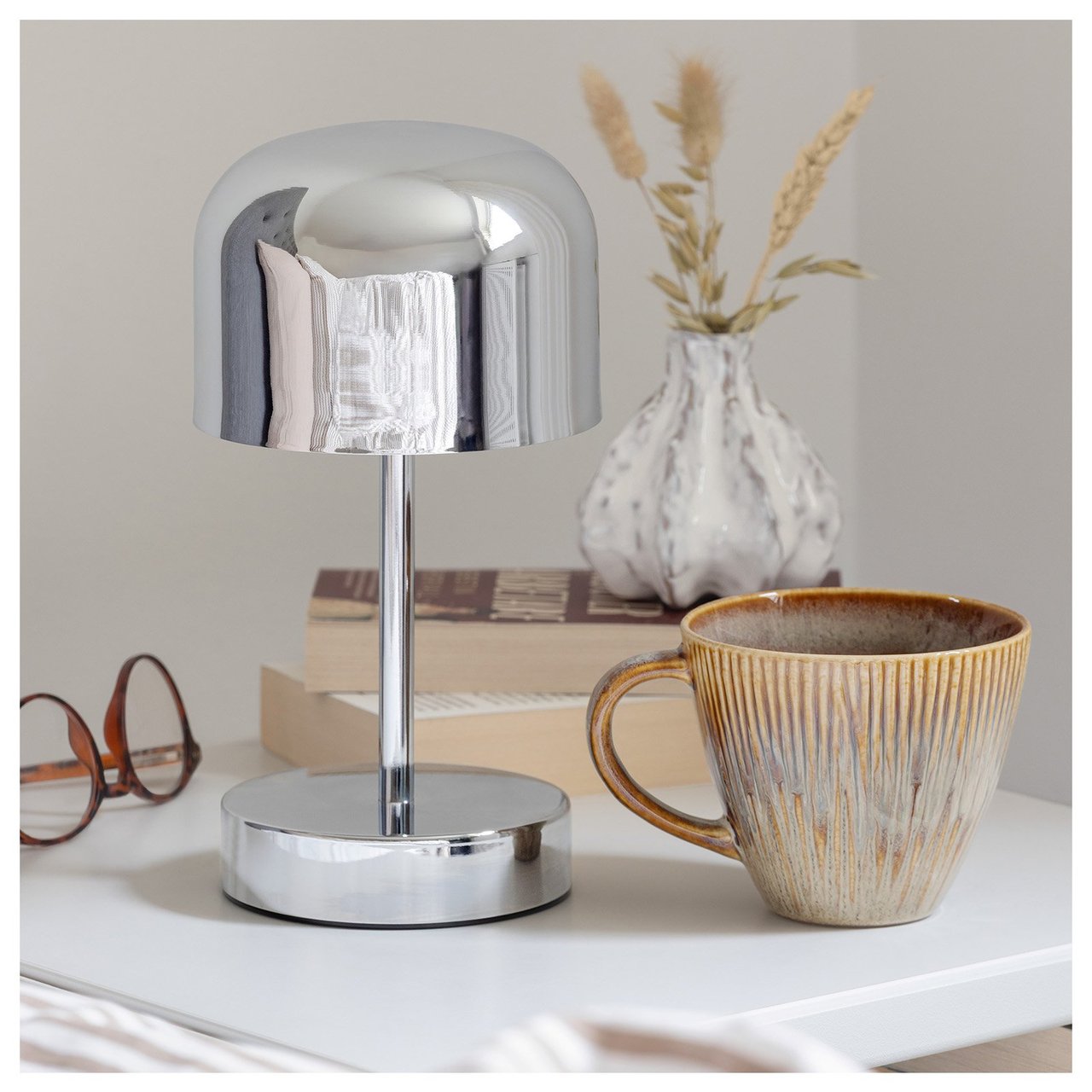 Eine verchromte Tischlampe auf einem Nachttisch, mit Tasse, kleiner Vase und Brille.