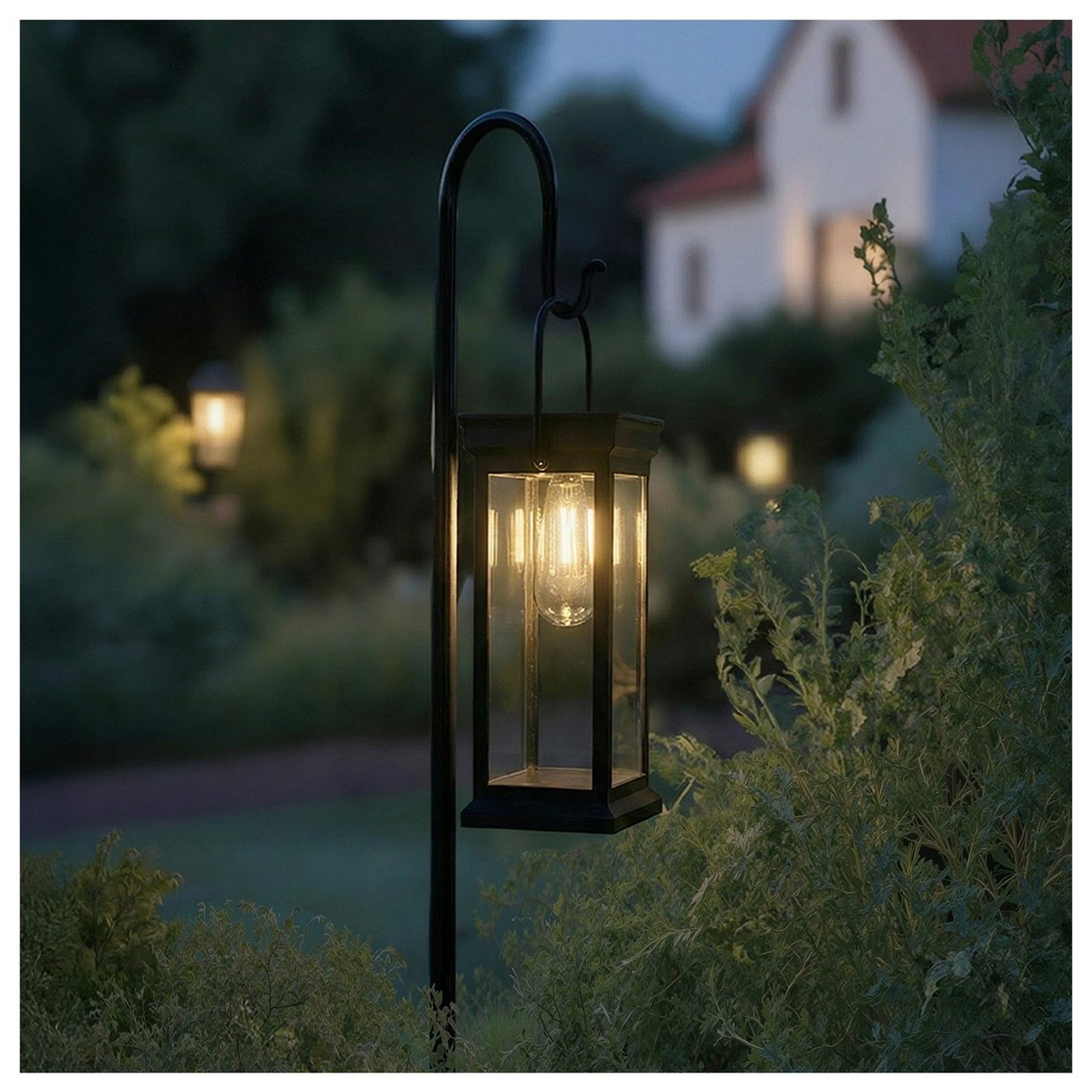 Liten svart solcellelykt henger i et svart metallstativ. I bakgrunnen ses et hvitt hus.