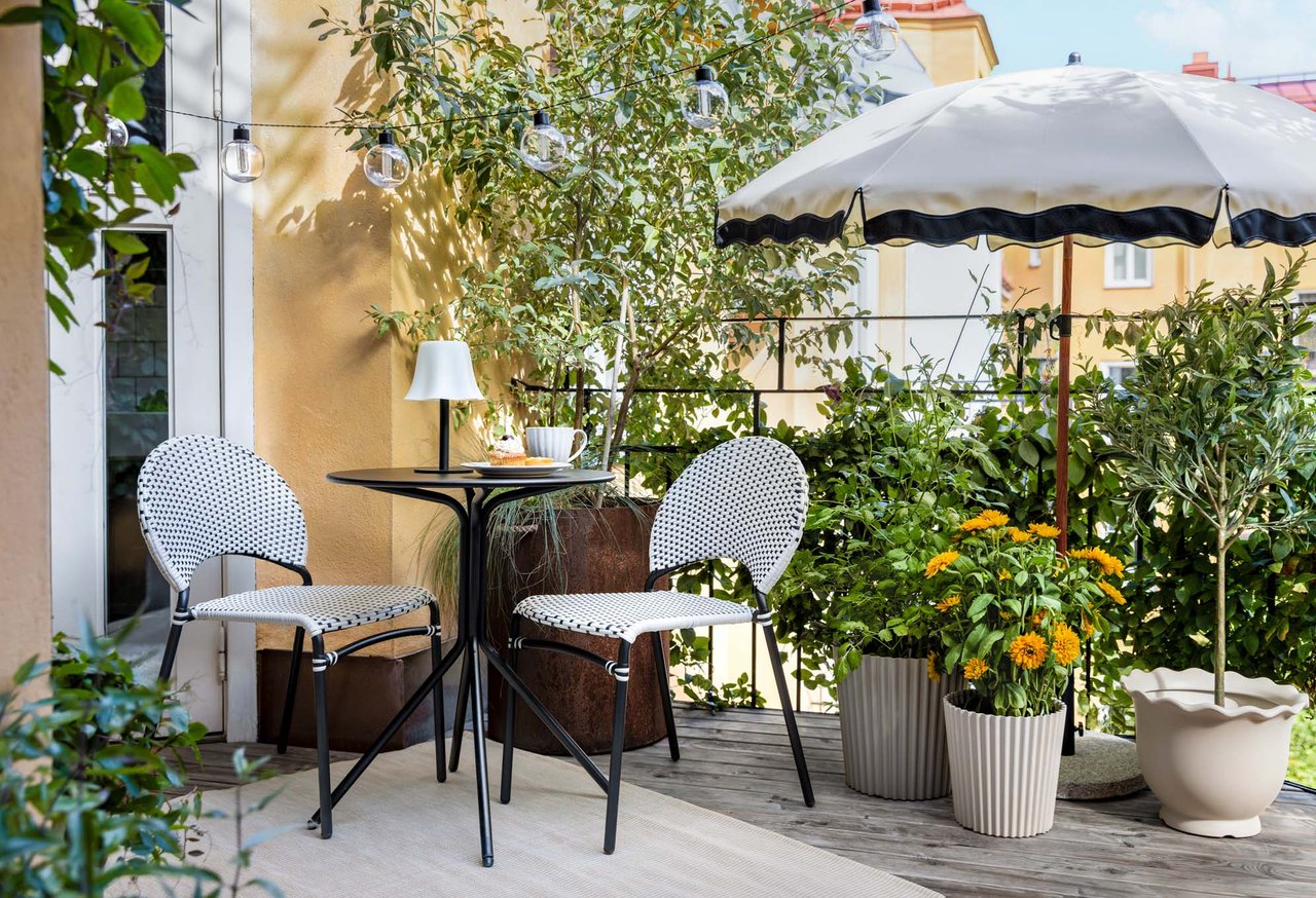 Sonniger Balkon mit kleinem Cafétisch, zwei Stühlen, Sonnenschirm, Lichterkette und vielen grünen Pflanzen.
