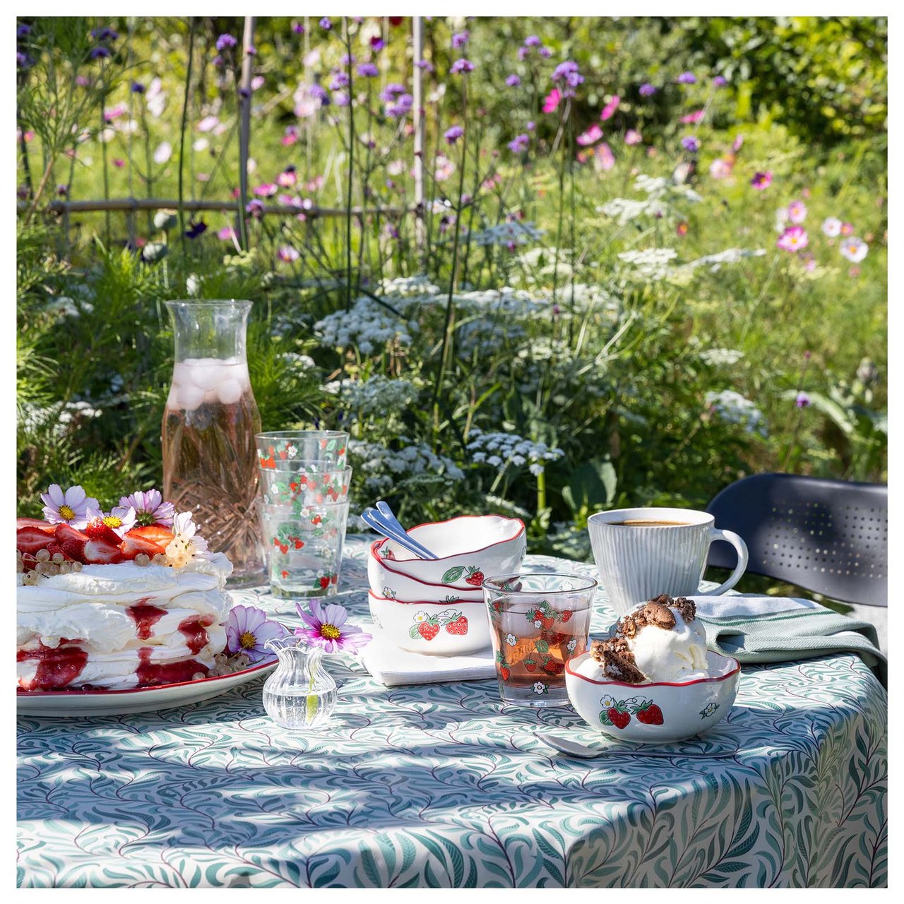 Ett dukat bord med jordgubbstårta och jordgubbsmönstrade skålar i en trädgård.