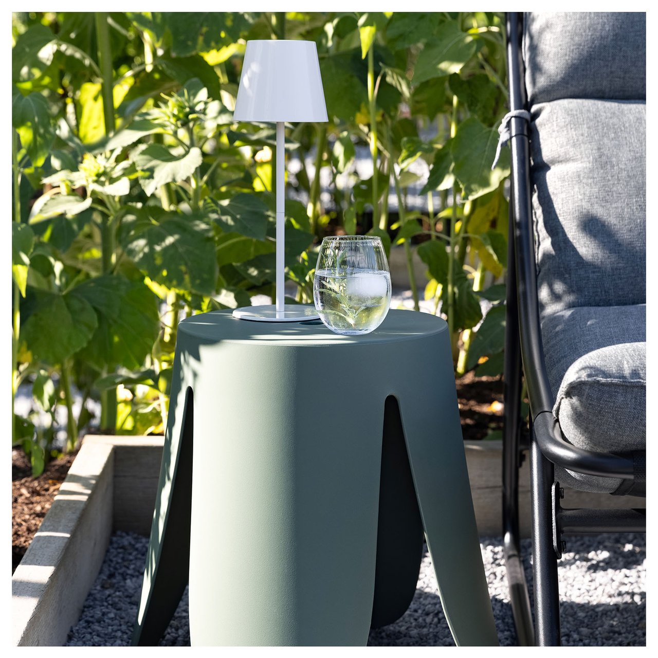 Grön pall med ett vattenglas och vit liten lampa står på en uteplats.